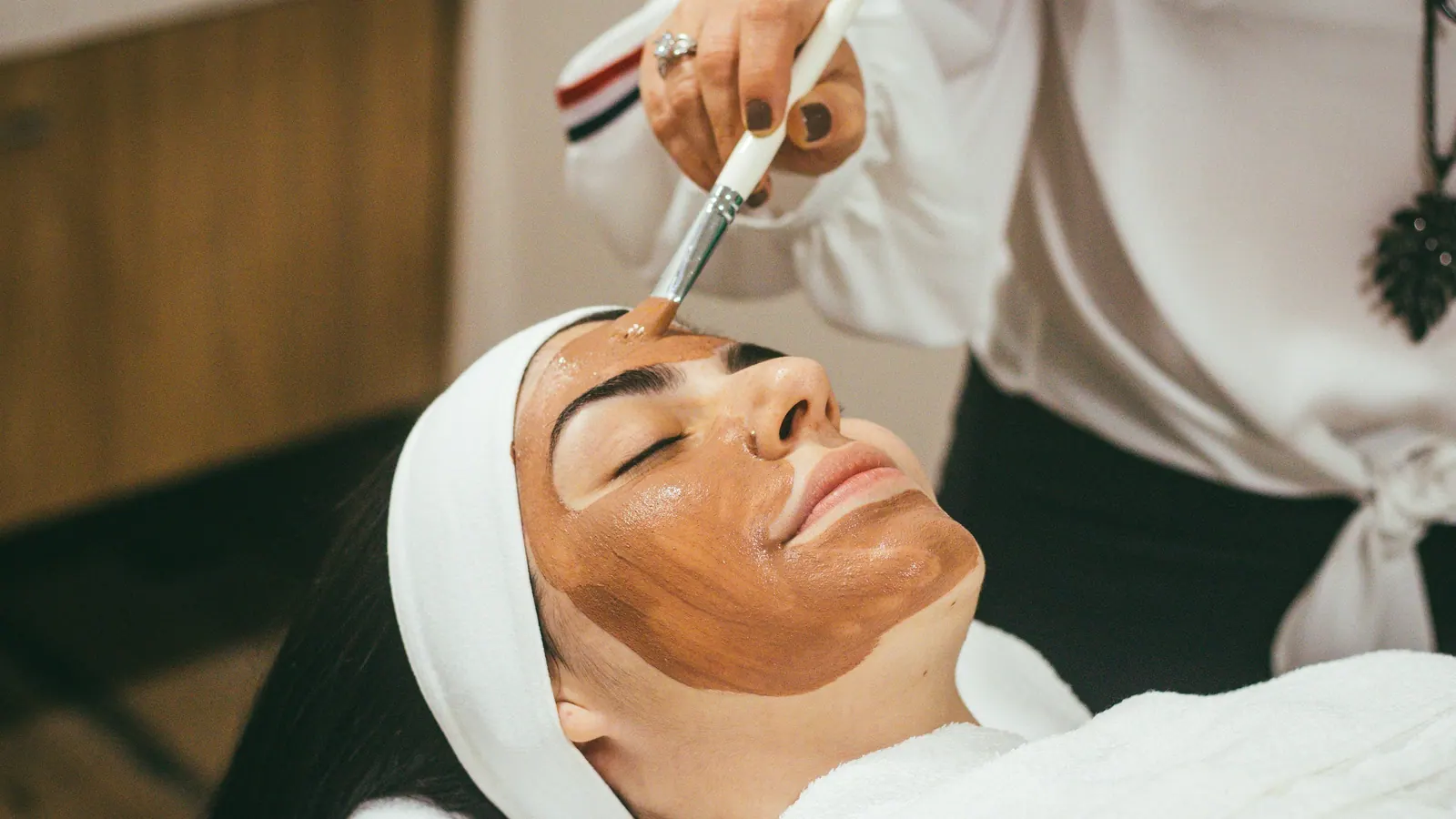 Esteticista aplicando máscara de argila no rosto de uma cliente deitada na maca